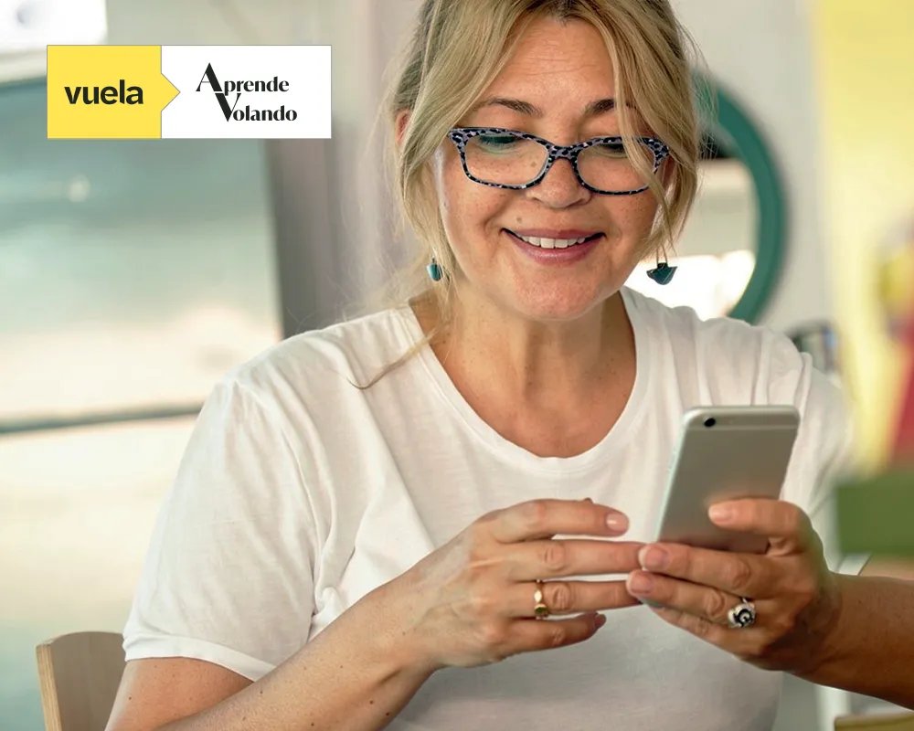 Mujer adulta con gafas utilizando un teléfono móvil y sonriendo, junto al logotipo de Vuela Aprende Volando en la esquina superior izquierda.