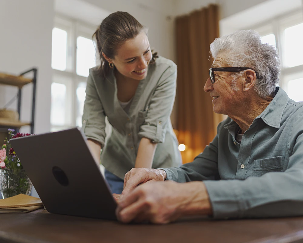 Mujer joven ayudando a un hombre mayor a utilizar un ordenador portátil en un entorno doméstico, ambos sonrientes frente a la pantalla.