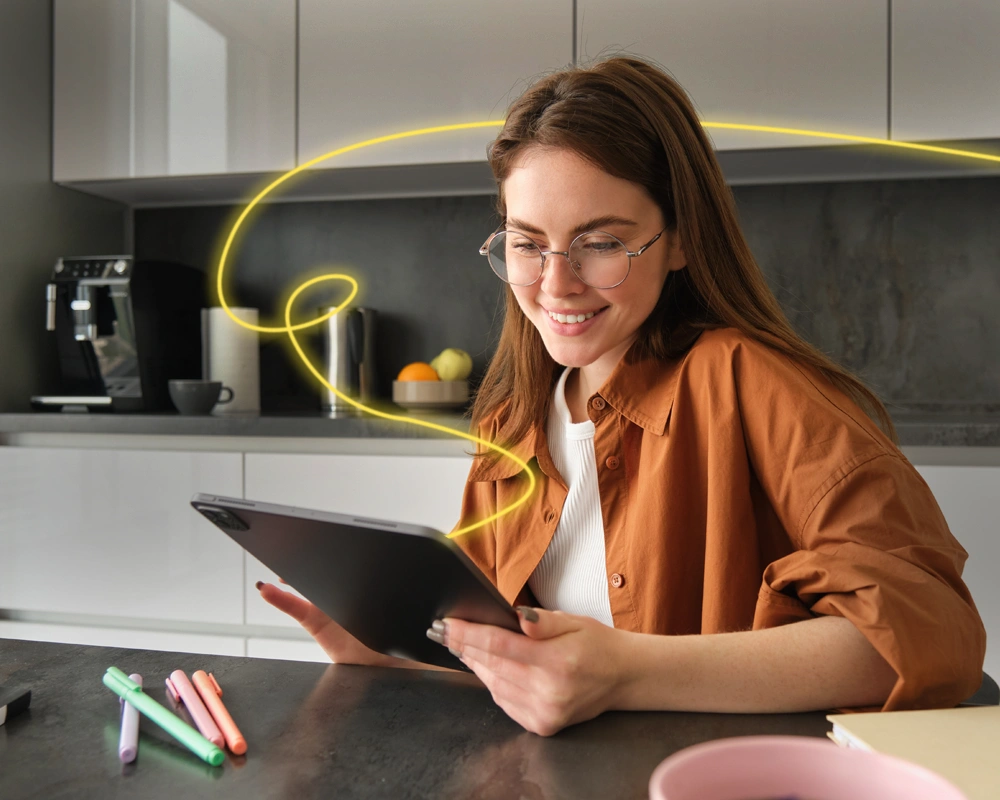 Mujer joven utilizando una tableta digital en una cocina moderna, con una línea luminosa amarilla que simboliza conexión o actividad digital a su alrededor.
