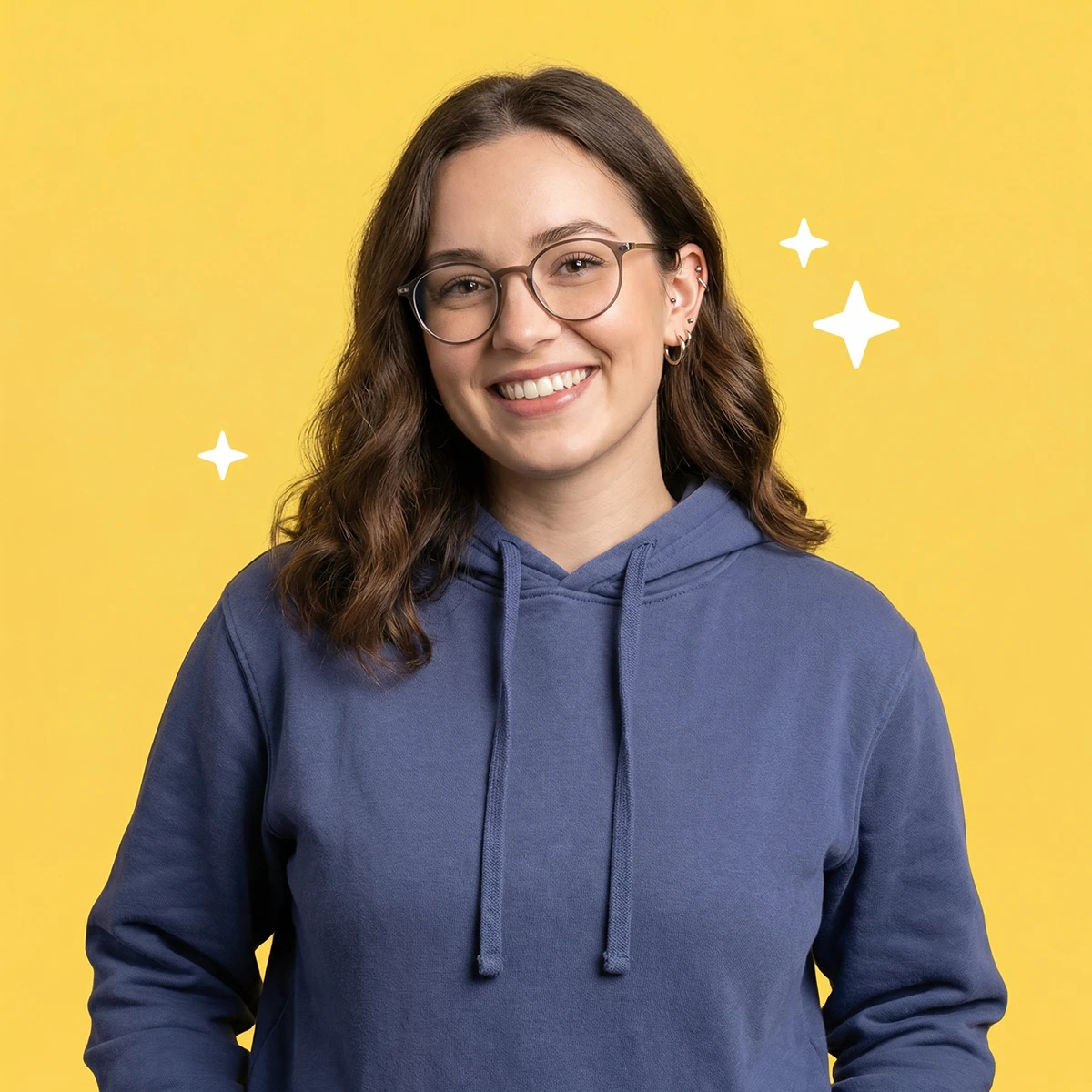 Retrato de una mujer joven con gafas y sudadera azul sonriendo frente a un fondo amarillo con pequeñas estrellas decorativas.