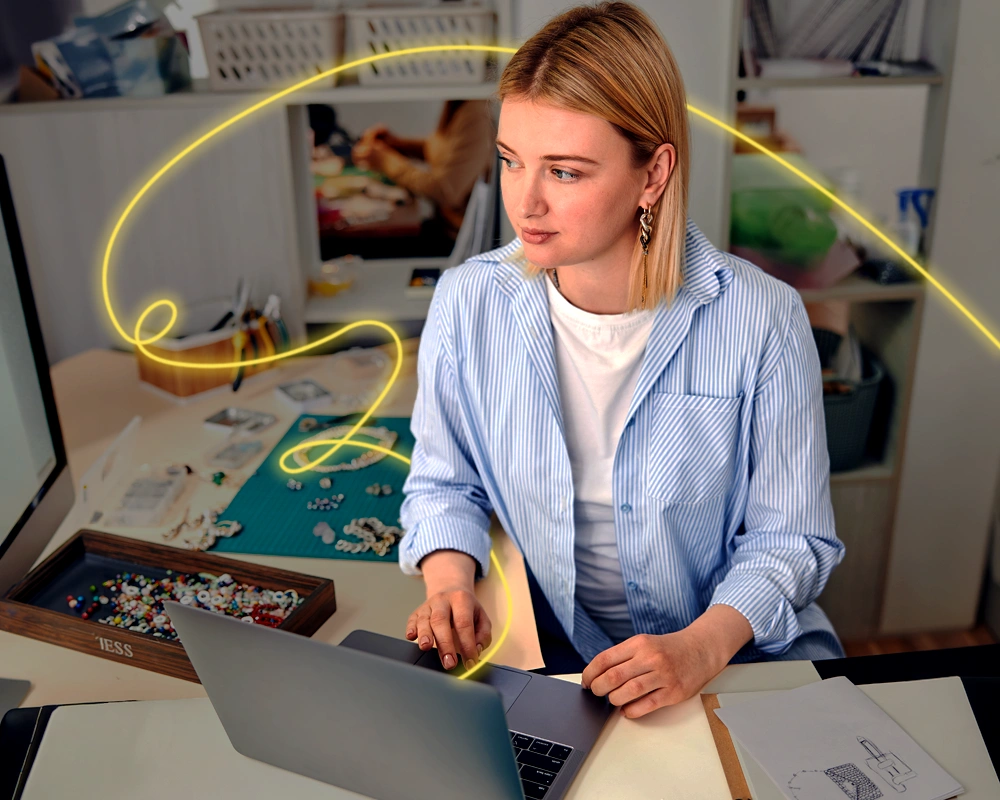 Mujer trabajando con un ordenador portátil en un taller creativo con materiales de bisutería sobre la mesa, mientras una línea luminosa amarilla simboliza conexión o impulso digital a su alrededor.