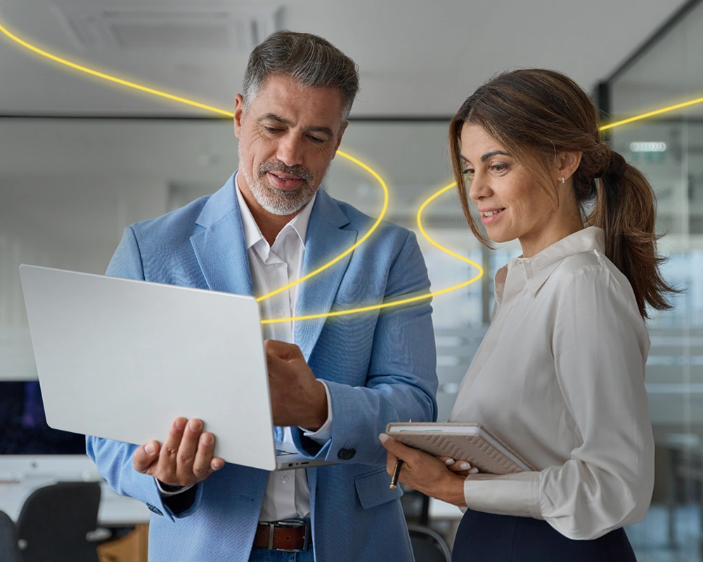 Dos profesionales en una oficina moderna revisan información en un ordenador portátil, mientras una línea luminosa amarilla simboliza la conexión o transformación digital.