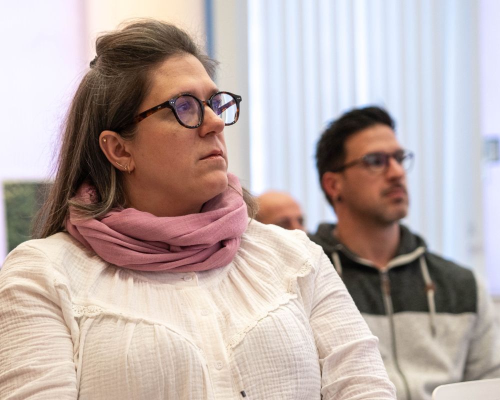 Mujer con gafas atendiendo en clase, detrás se observa otro alumno con gafas atendiendo.